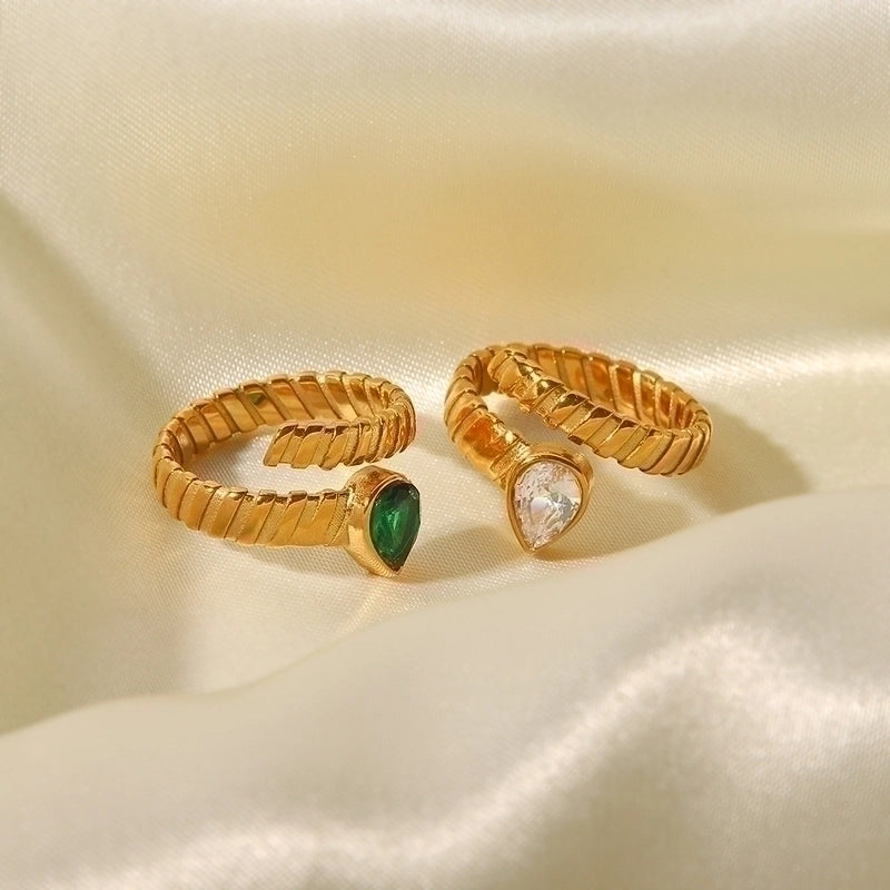 Two gold adjustable rings with a ridged band on fabric. One ring has a green teardrop stone, the other has a clear, diamond-like teardrop stone.